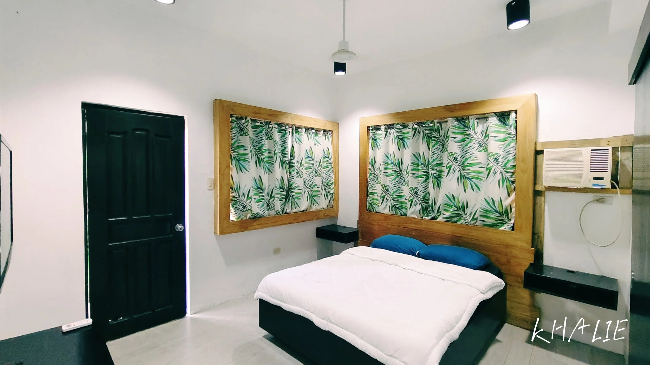 A bedroom with white walls, black door, two picture windows with tropical leaf curtains, a floating black shelf, a bed with white bedding, blue pillows, a wall-mounted air conditioning unit, and ceiling lights.