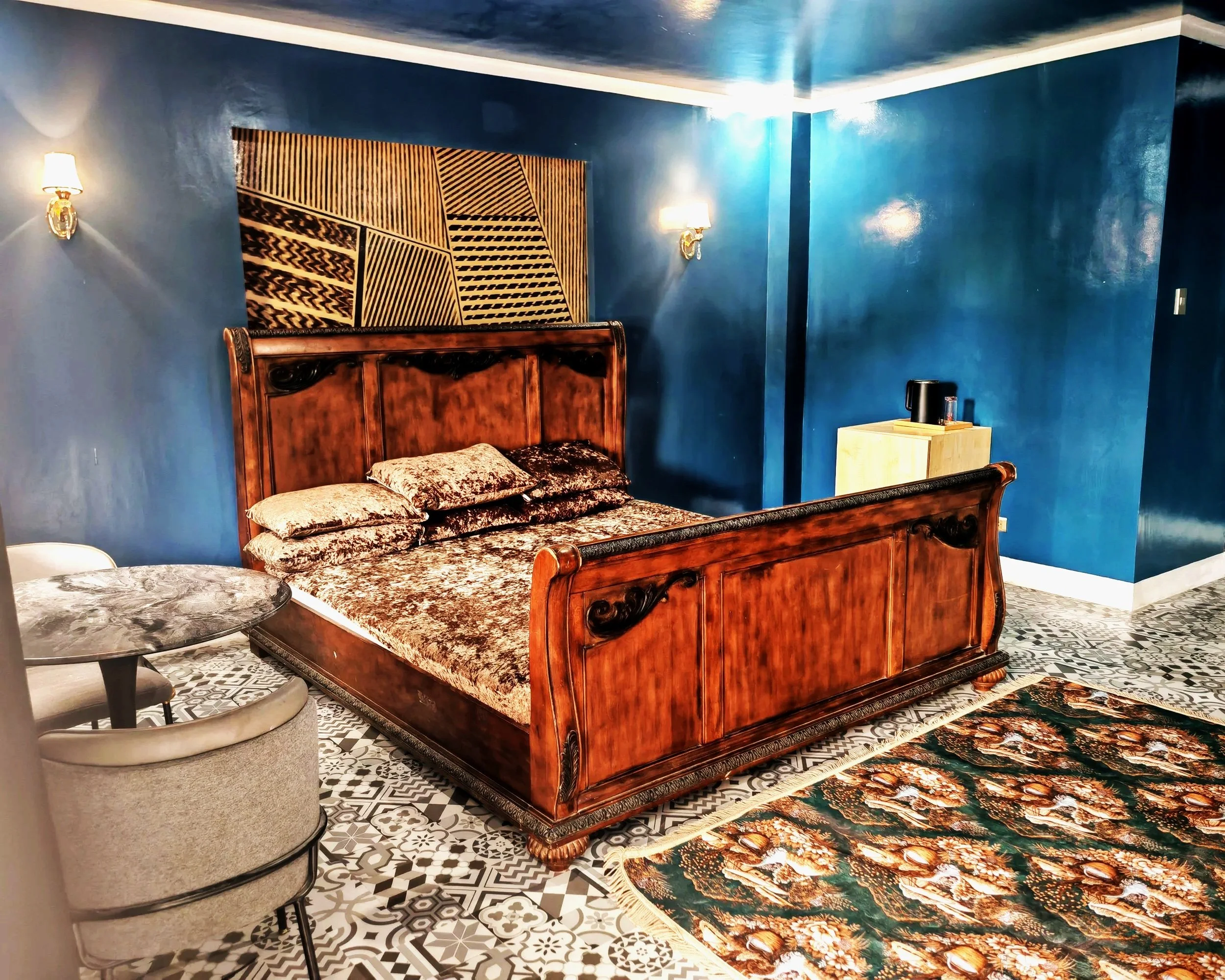 A bedroom with blue painted walls, a large wooden bed with carved details, patterned bedding, a decorative tapestry above the bed, two wall-mounted lamps, a small wooden side table with a black kettle, a patterned rug, and tiled flooring.