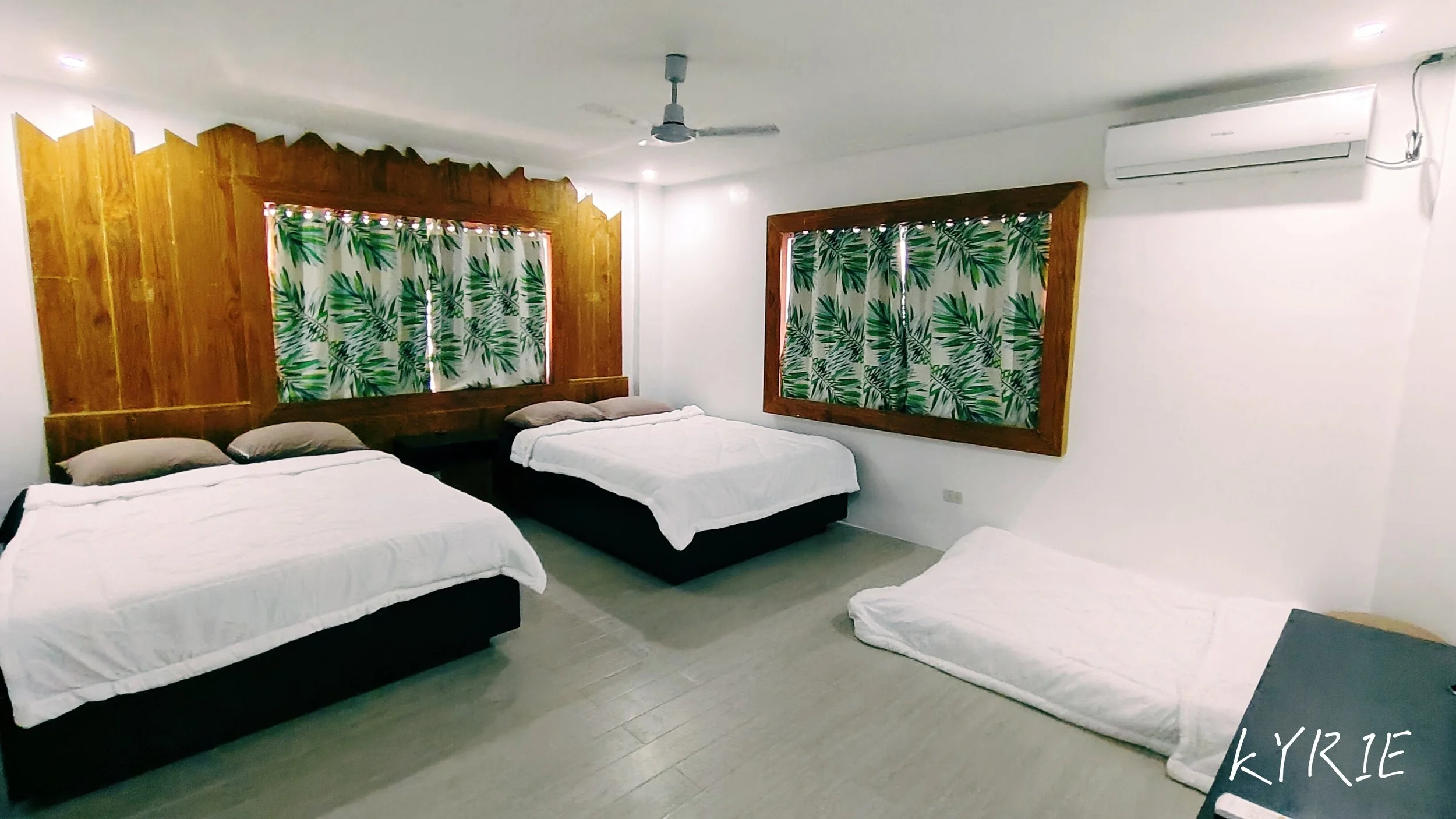 A bedroom with four beds, a wooden headboard, patterned curtains with green leaves, a ceiling fan, and an air conditioning unit on the wall.