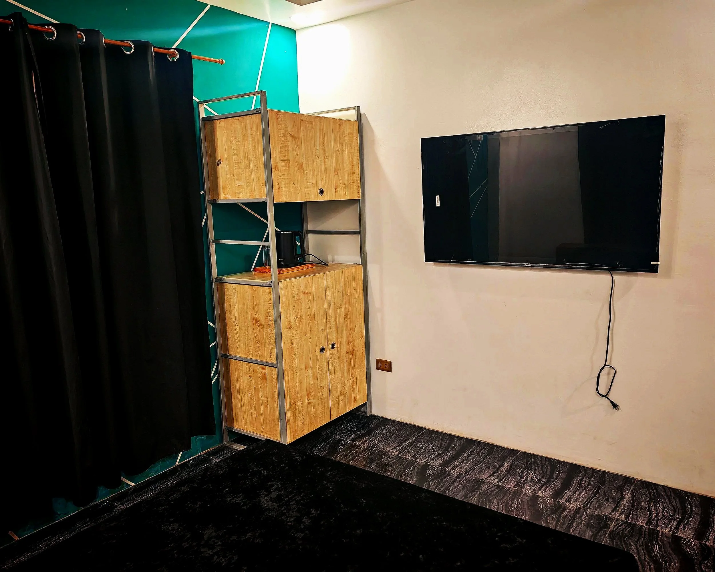 Room with black curtains, a wooden shelf, a large flat-screen TV, and a power cord on the wall.