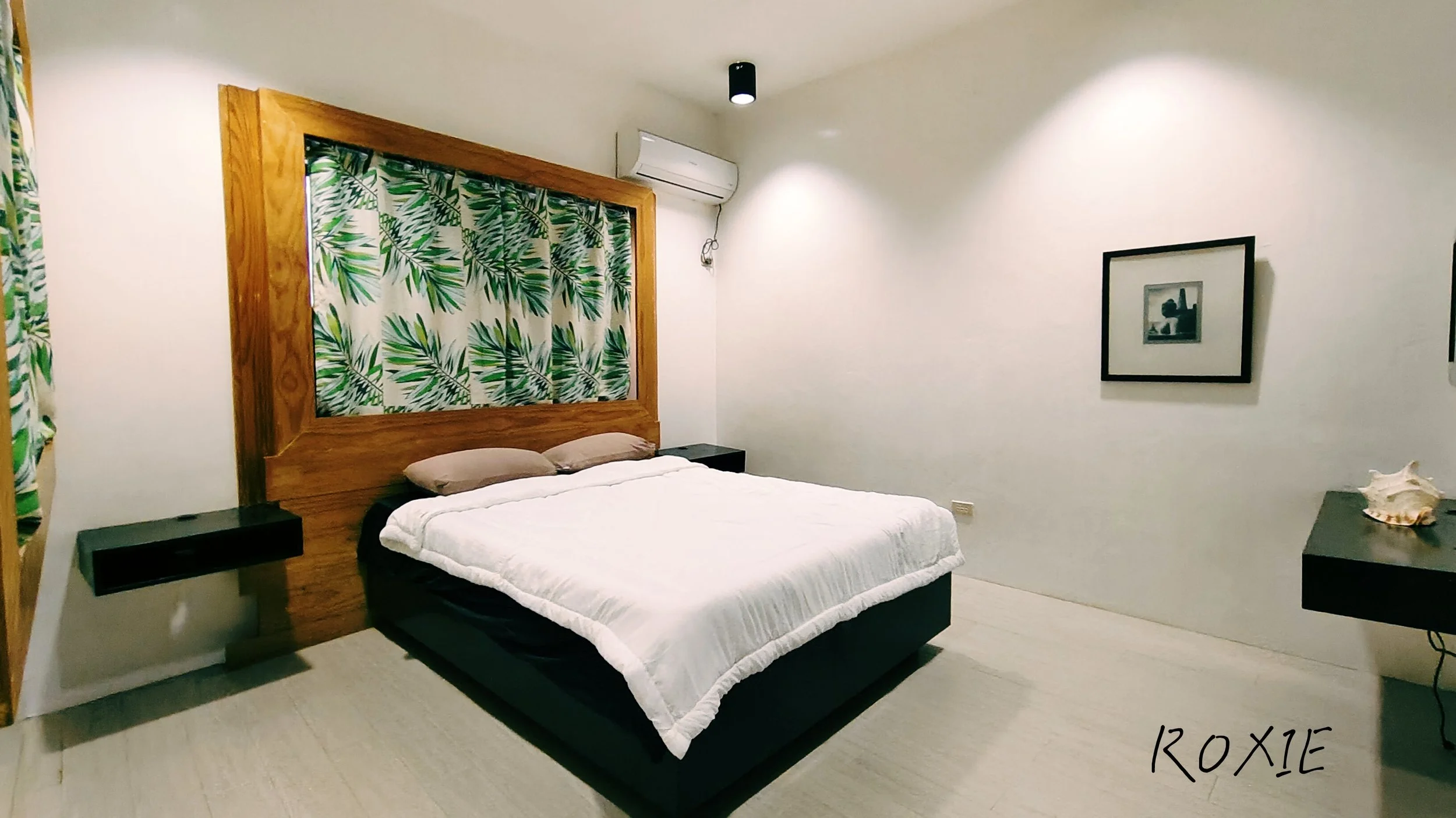 A bedroom with a double bed, white bedding, and two beige pillows. The headboard is made of wood with a tropical leaf pattern on the fabric above it. There are black bedside tables on each side, and a framed black-and-white photo on the wall. An air conditioning unit is mounted on the wall, and a small black ceiling light is visible. The floor is light-colored, and there is a piece of furniture with a seashell on top in the corner.
