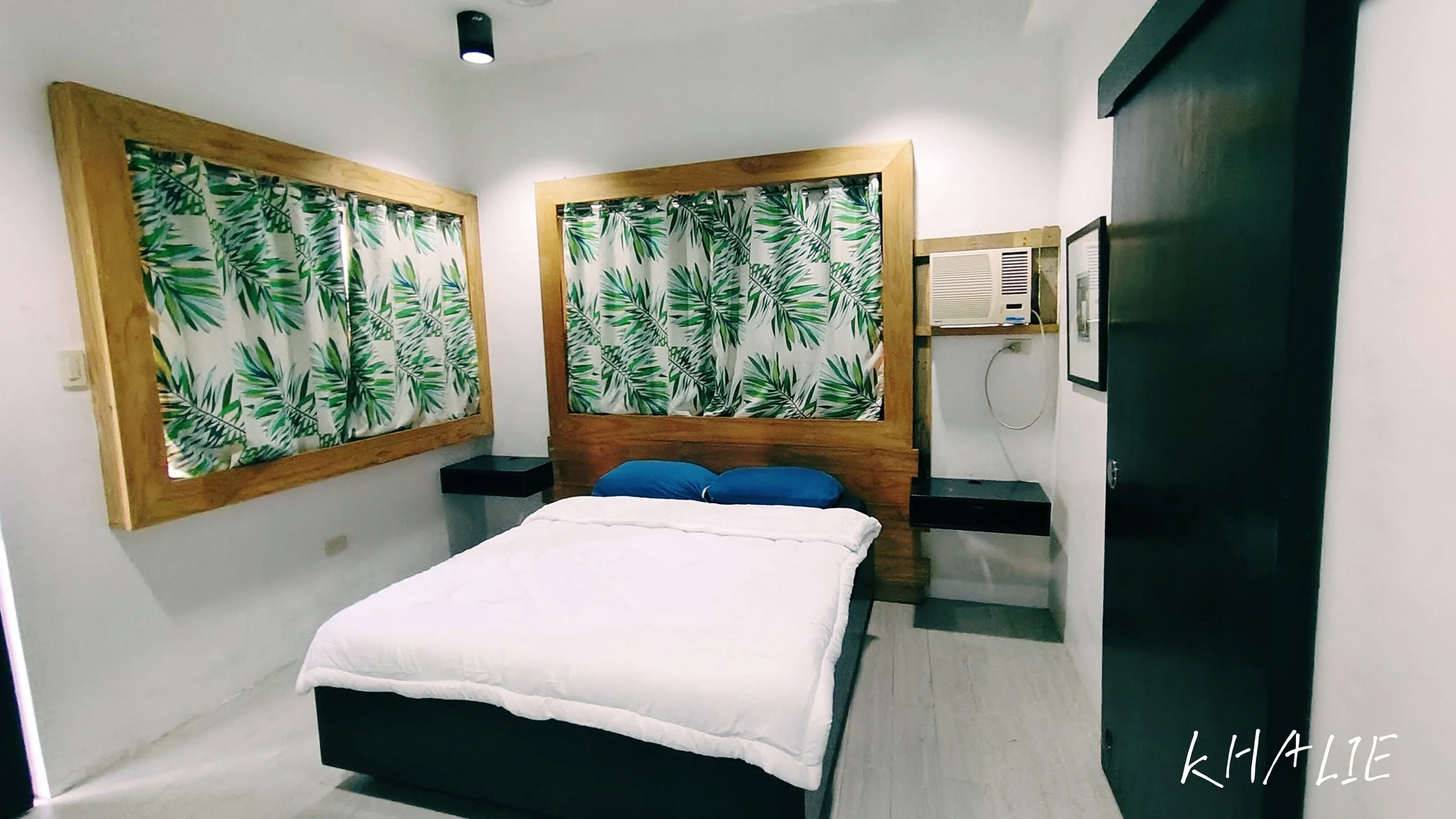 A simple bedroom with a white bed, blue pillows, and tropical print curtains covering two windows. There are black nightstands on either side of the bed, with an air conditioning unit mounted on the wall to the right. The room has white walls and a black ceiling light.