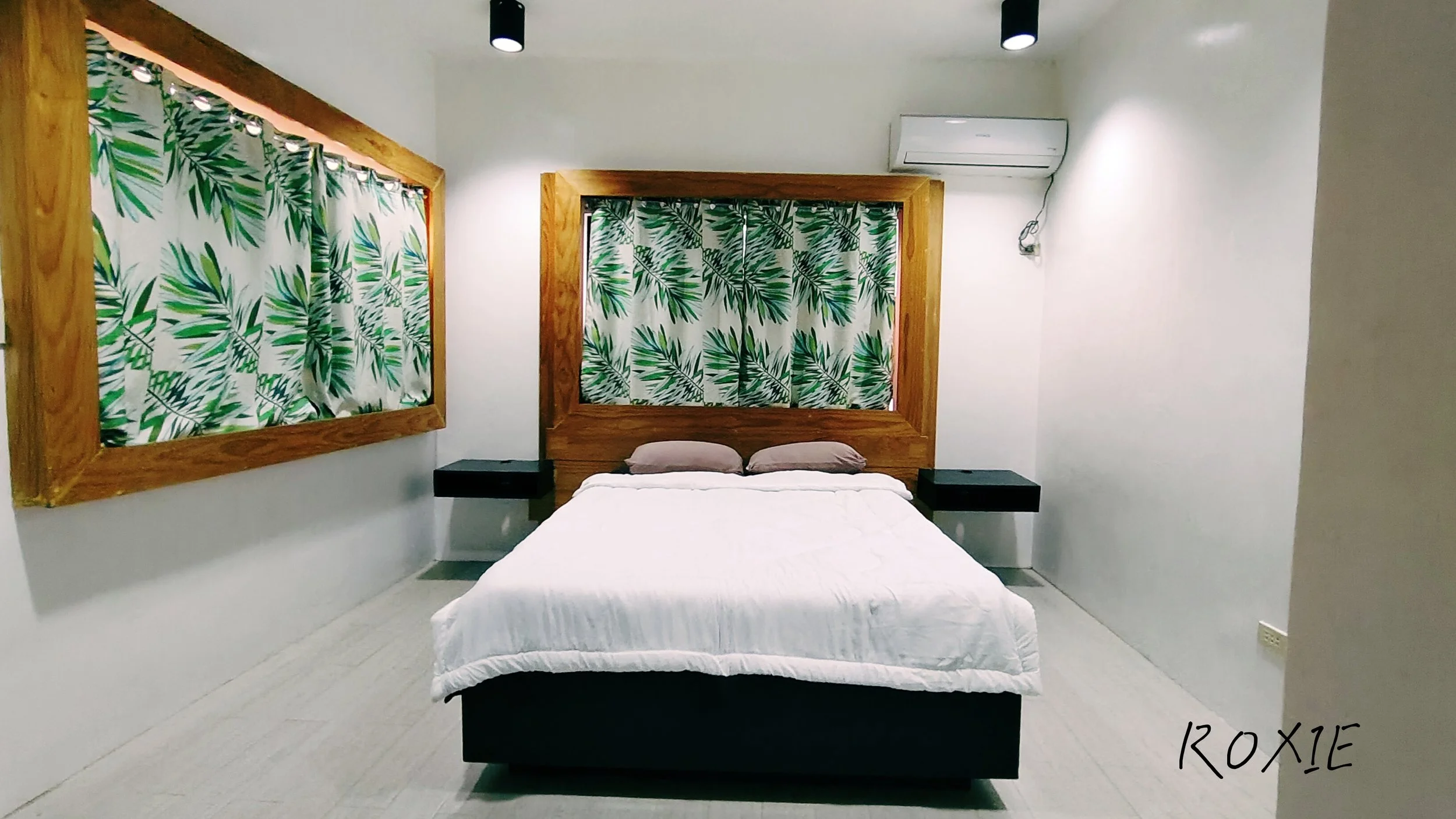 A bedroom with a white bed, two brown pillows, and a wooden headboard. There are two large windows with green and white tropical leaf patterned curtains, framed with wood. The walls are white, and there is a split air conditioner mounted on the wall. Two black ceiling lights are visible, and a black nightstand is placed beside the bed. An inscription in the lower right corner reads '12x15'.