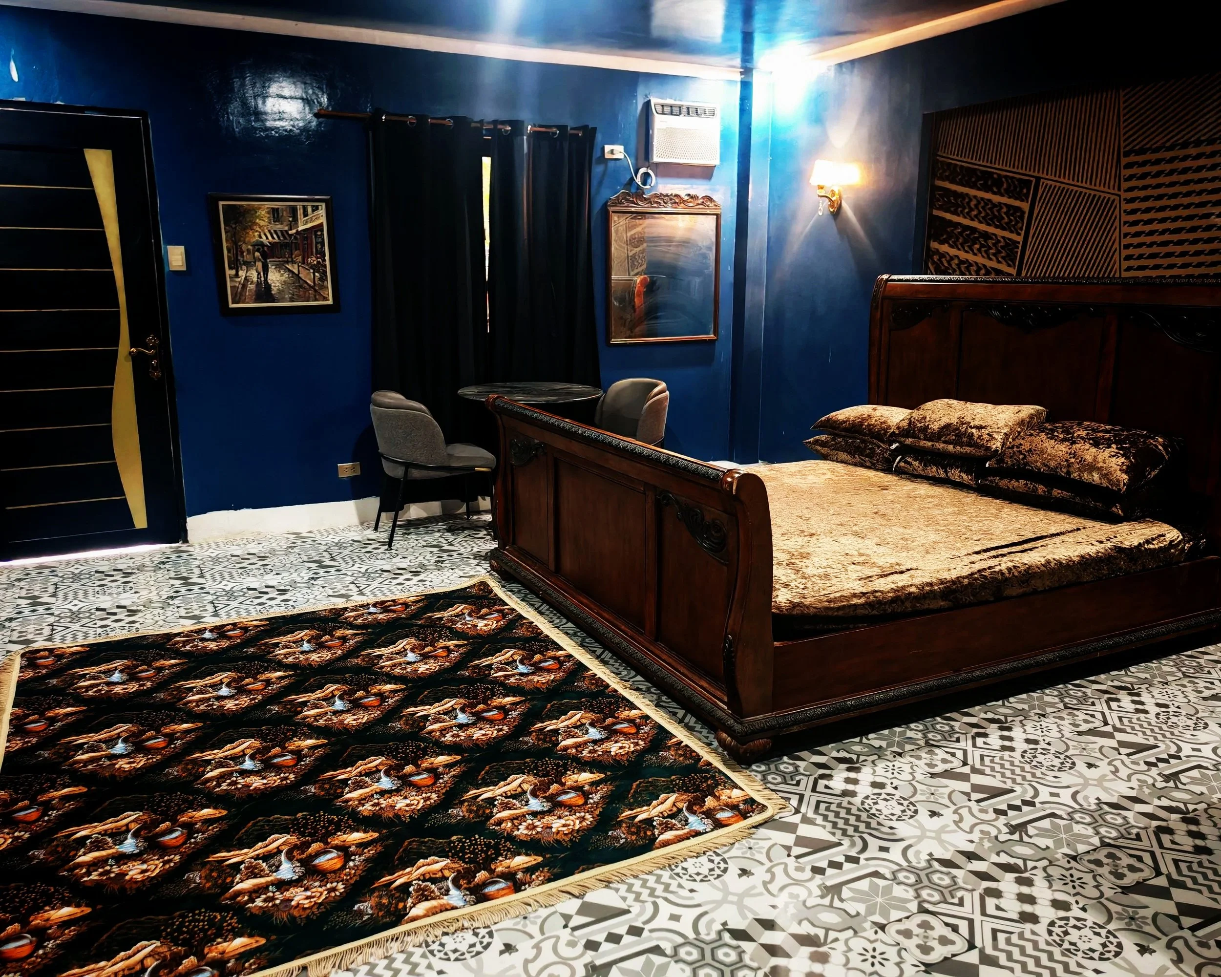 A bedroom with dark blue walls, patterned tile flooring, a wooden bed with floral bedding, a mirror, a small table with two chairs, and artwork on the walls.