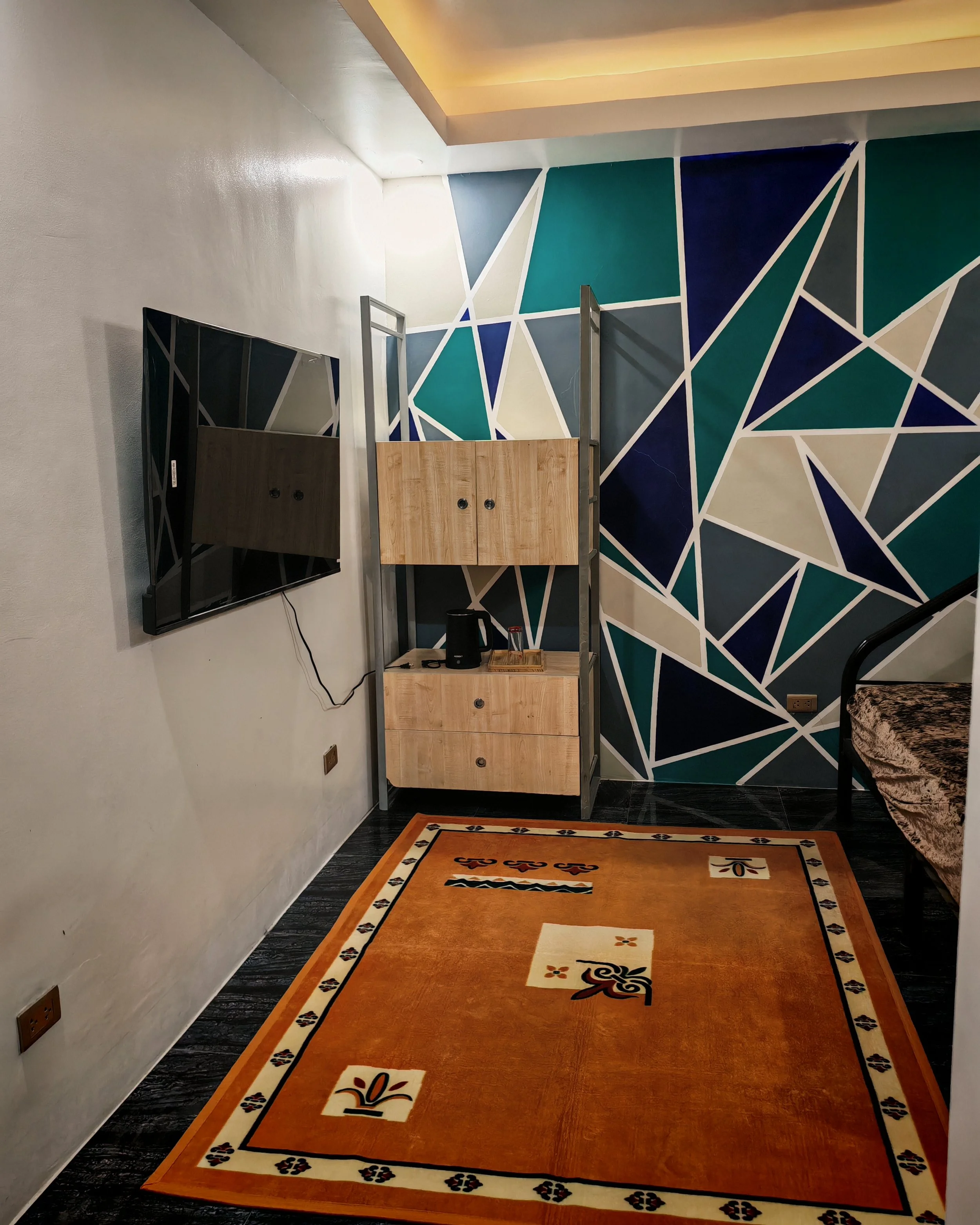 A hotel or lobby area with a large orange rectangular rug featuring black and white decorative patterns at the border and smaller patterns toward the center. To the left, a flat-screen television is mounted on the wall, with a power cord hanging down. To the right, a wooden cabinet with drawers and a black electric kettle on top. Behind the cabinet, a geometric, stained-glass style wall mural with shades of blue, teal, gray, white, and beige. Partially visible on the far right, a piece of furniture with a marble or faux-marble top.