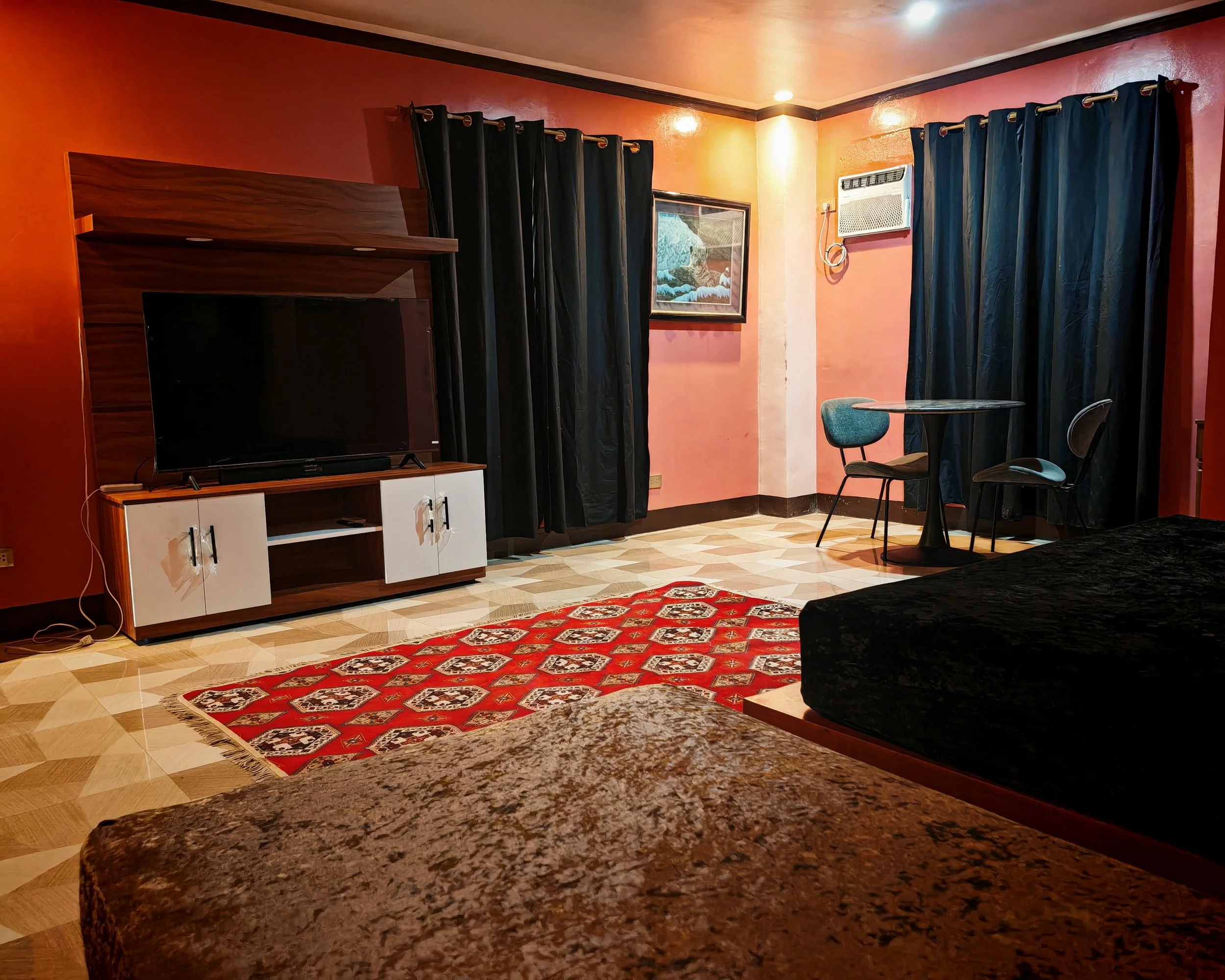 Interior of a room with pinkish-orange walls, black curtains, a wall-mounted air conditioner, a small round dining table with two chairs, a painting, a television on a wooden stand, and a red patterned rug on the wooden floor.