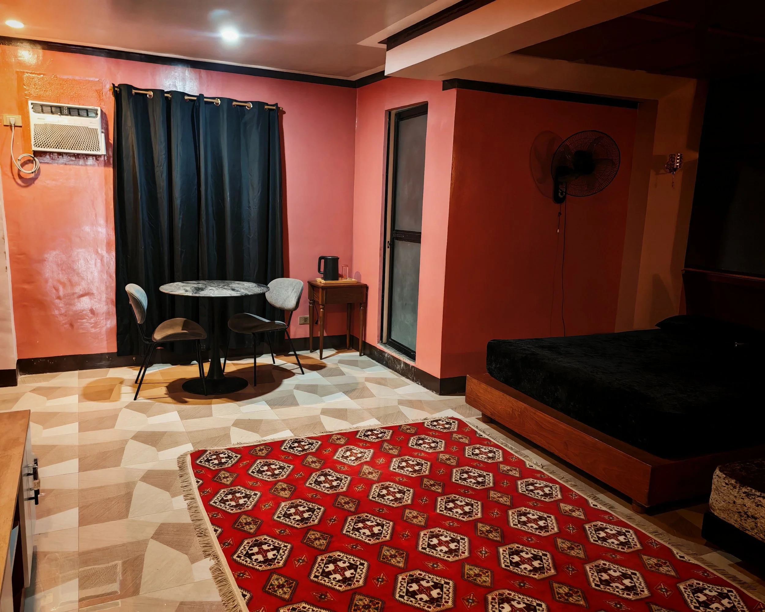 Living room with pink walls, black curtains, a small round table, four chairs, a red patterned rug, a window with a screen, a wall-mounted fan, and a wooden side table with a black thermos and glasses.