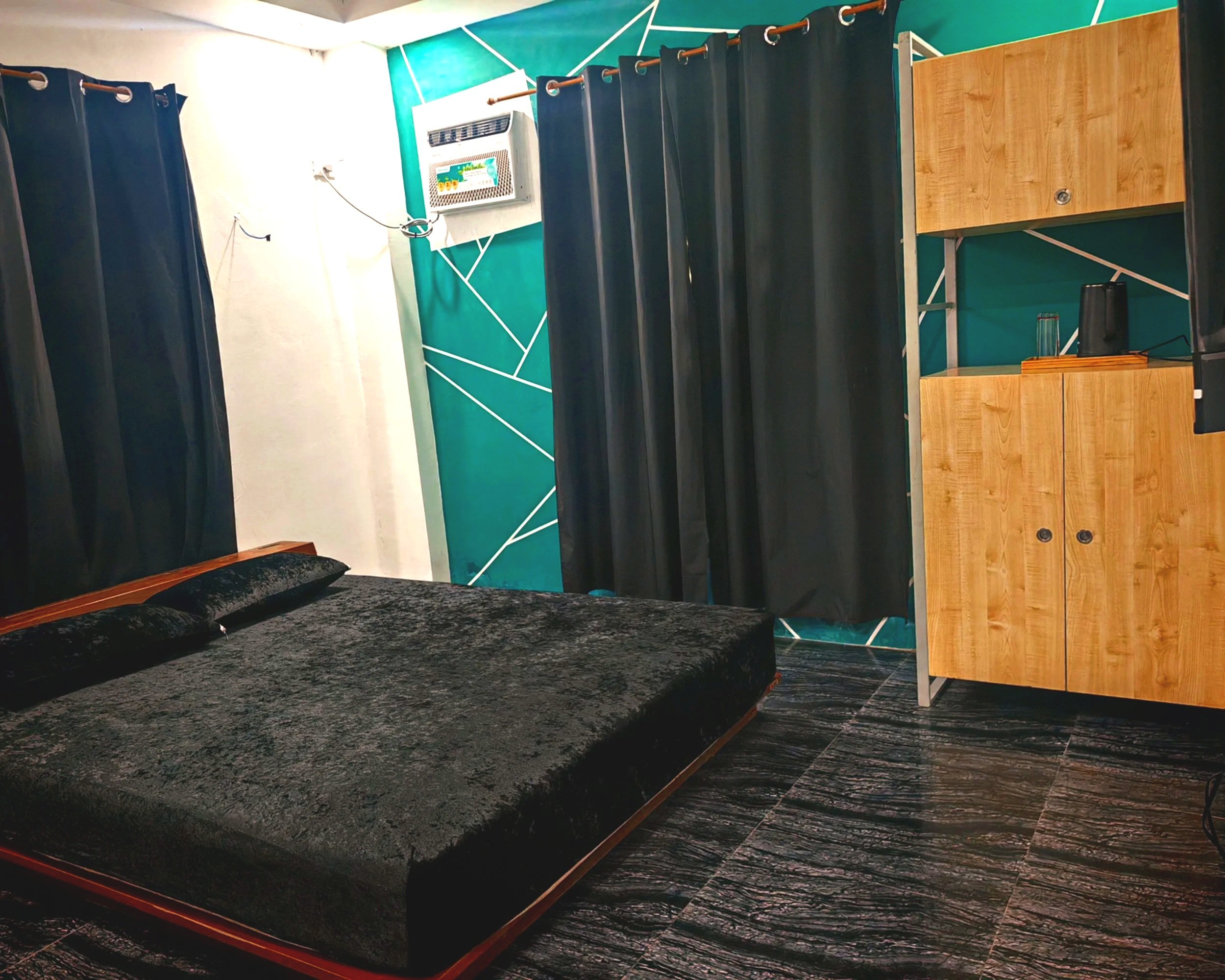A room with dark curtains, a wooden desk, and an HVAC unit on a teal wall with geometric white lines. The lower part of a black couch is visible in the foreground.