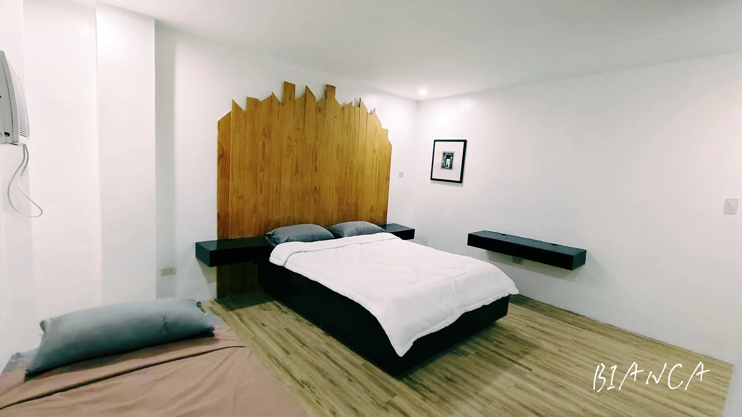 A bedroom with a large bed, two black nightstands, a wooden headboard, and two gray pillows. A framed black-and-white photo hangs on the white wall. The floor is wooden, and the room has minimal decor.