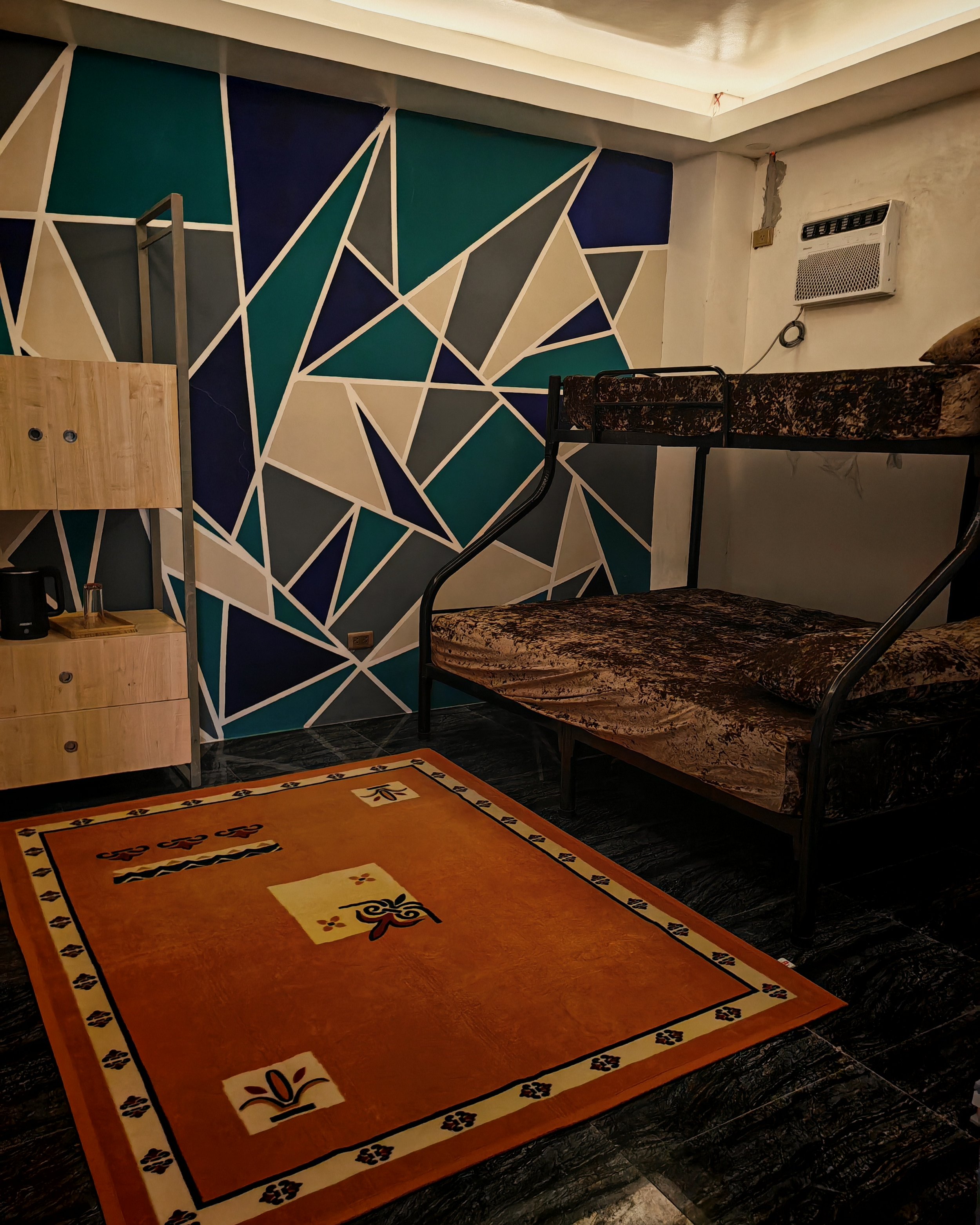A room with a geometric patterned wall, an air conditioner mounted on the wall, a brown velvet couch, a wooden shelf with a small glass of drink, and an orange area rug with black, white, and yellow patterns.