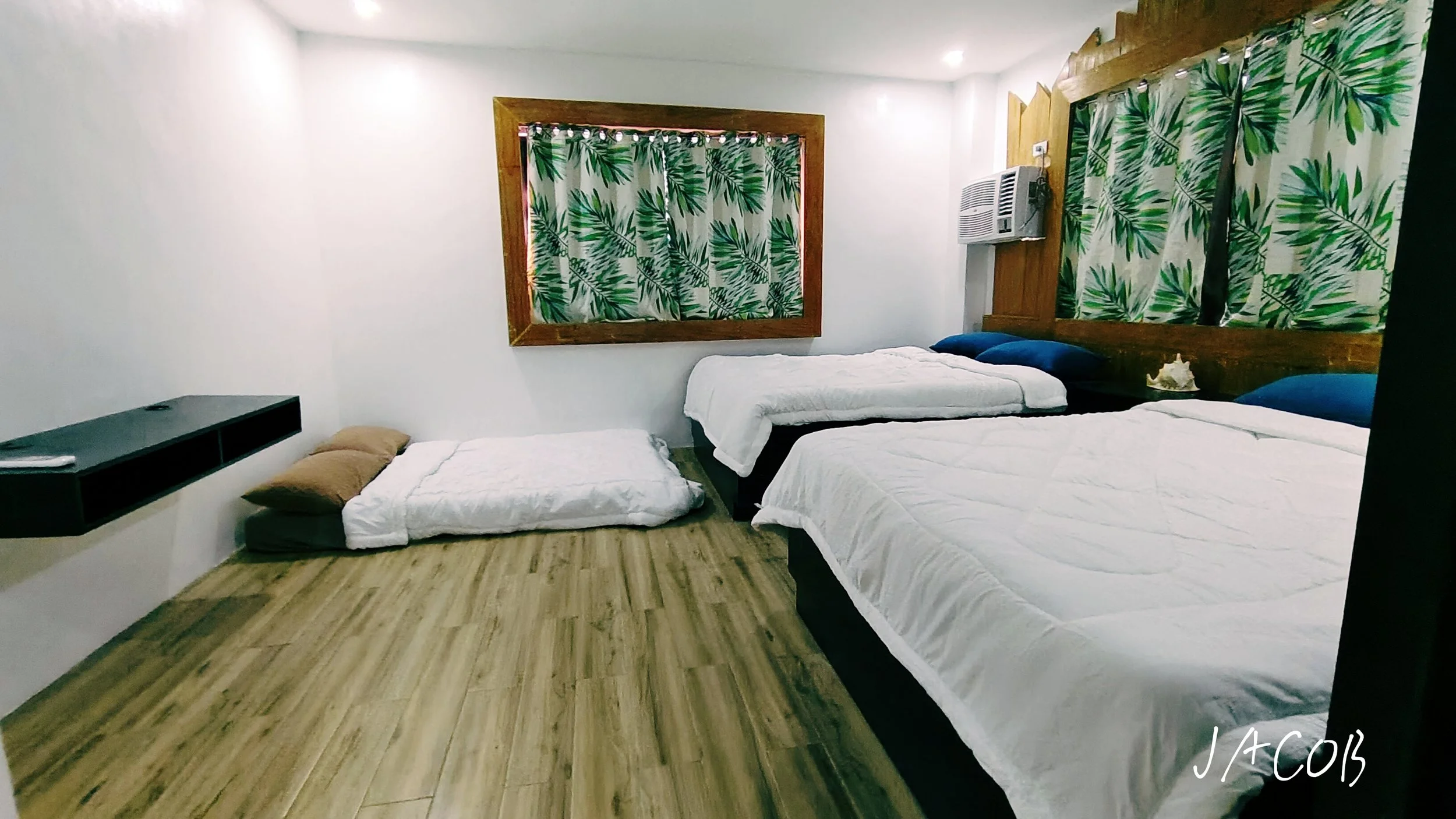 A bedroom with three beds, two with dark blue pillows and one on the floor with tan pillows, wooden floors, and tropical leaf-patterned curtains covering windows. There is a small dark shelf on the left wall and an air conditioning unit above the middle bed.