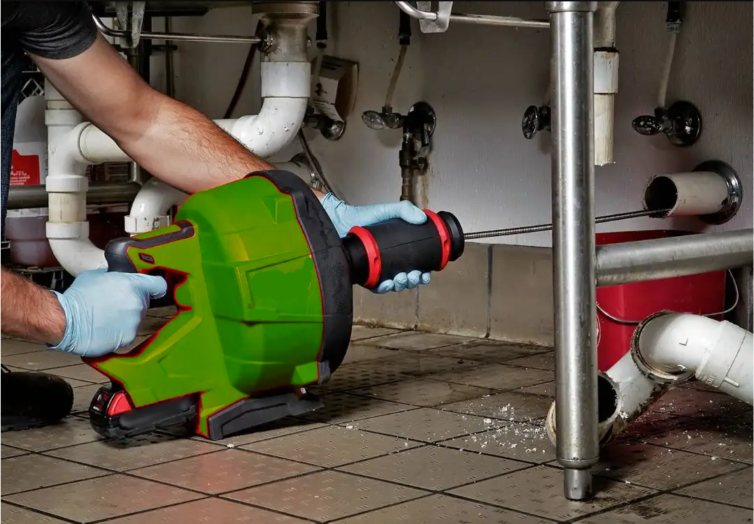 Person using a cordless drain auger to remove a blockage from under a sink in a plumbing area.