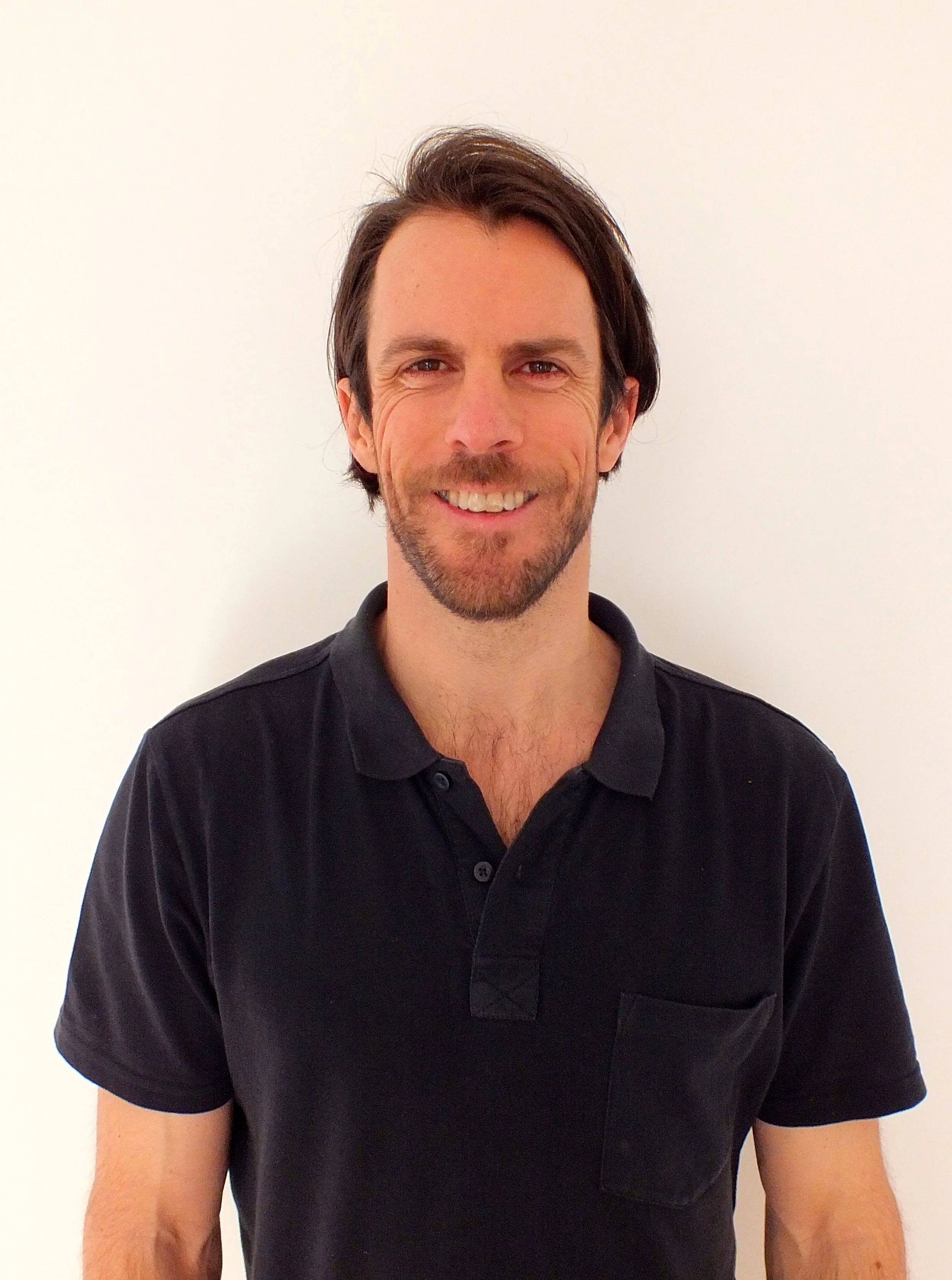 A smiling man with brown hair, facial hair, and light skin, wearing a black polo shirt, standing against a plain white wall.