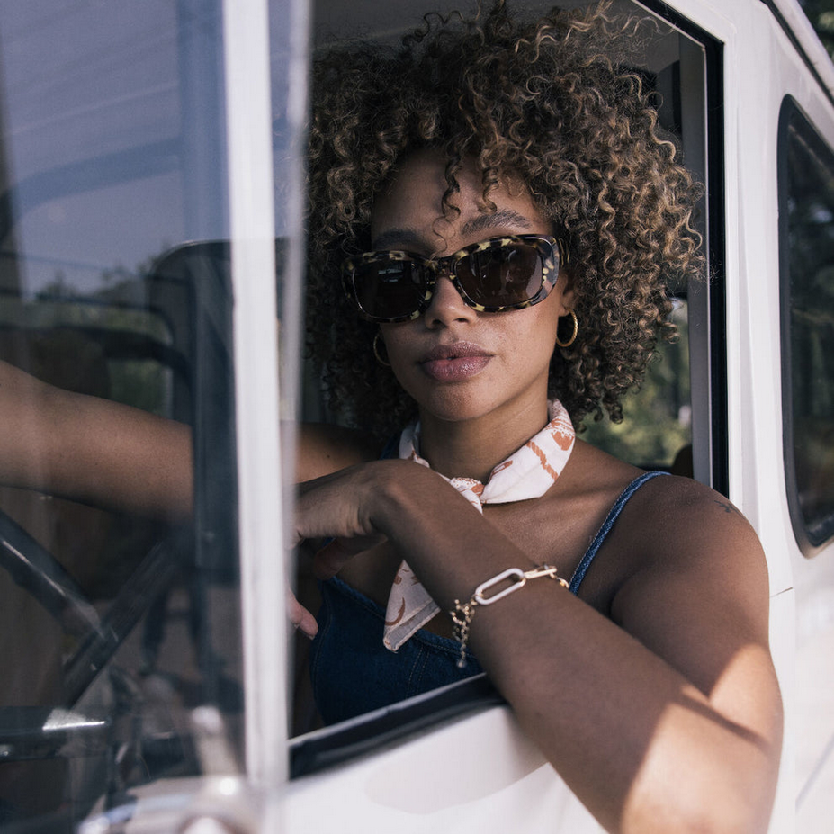 Woman with curly hair wearing sunglasses, a patterned scarf around her neck, and a denim sleeveless top, sitting in the driver's seat of a vehicle.
