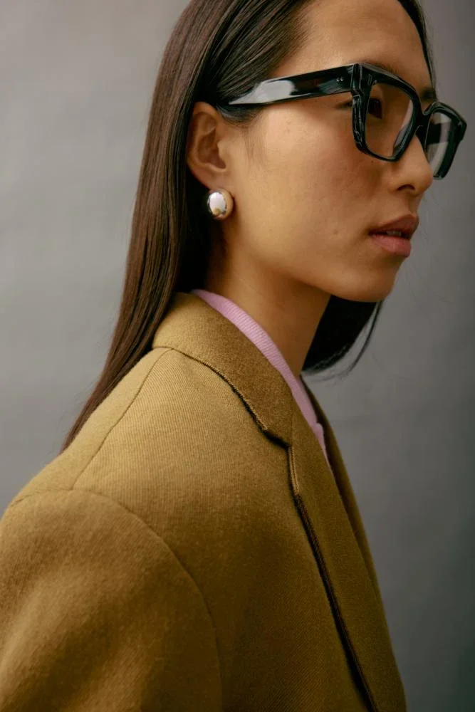 Side profile of a woman wearing black glasses, pearl earrings, a beige blazer, and a pink shirt underneath, against a plain gray background.