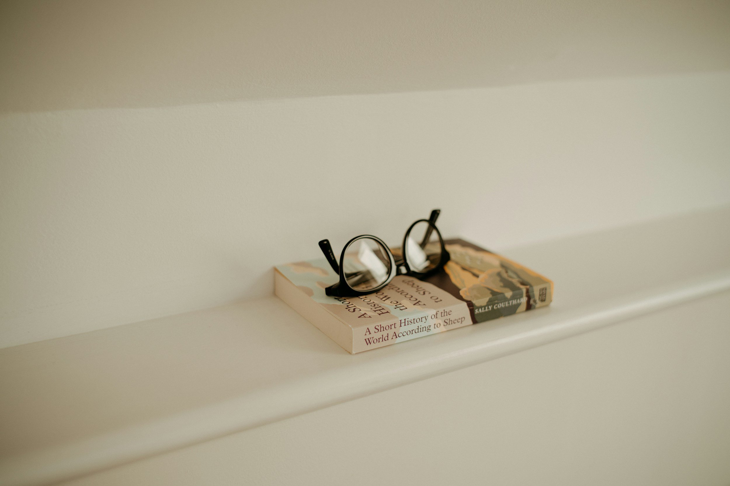 A pair of black eyeglasses resting on top of a book titled 'A Short History of the World According to Sheep' by Sally Coulthard, placed on a white shelf against a plain beige wall.