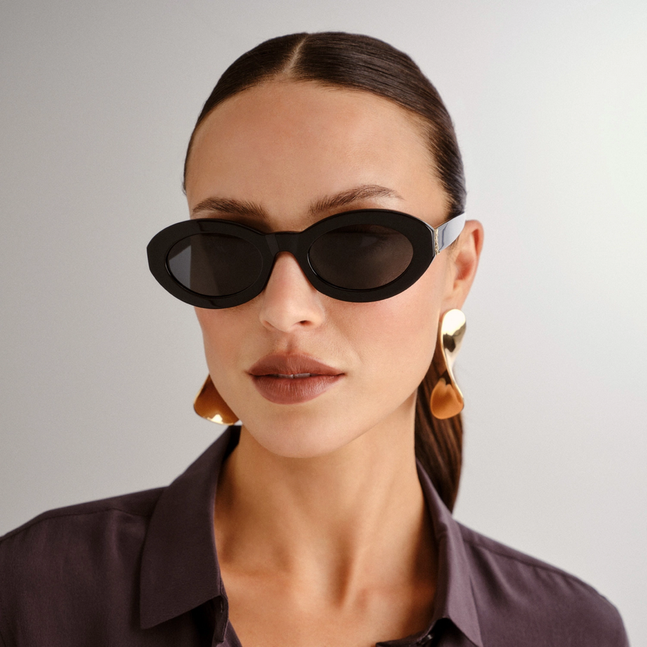 Woman with brown hair, wearing black sunglasses with oval lenses, large gold earrings, and a dark blouse against a plain background.