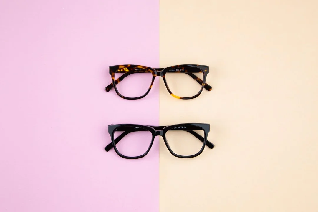 Two pairs of eyeglasses, one with tortoiseshell framing and one with black framing, are positioned horizontally on a split pink and cream background.
