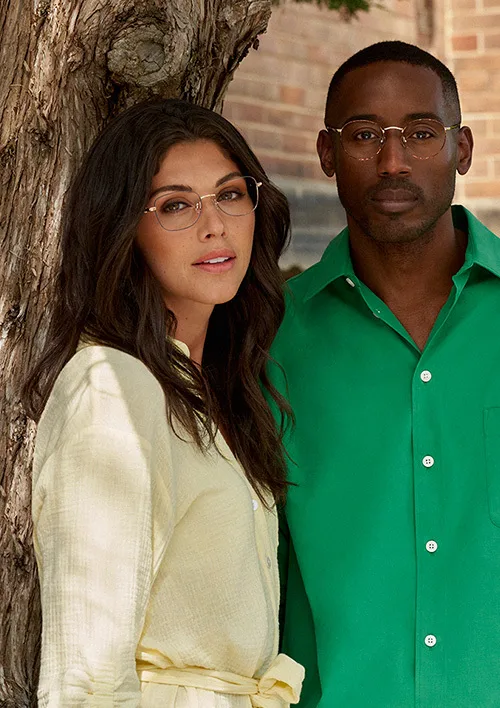 A woman and a man standing outdoors next to a tree, looking at the camera. The woman has long dark hair and glasses, wearing a light-colored outfit. The man has short hair, glasses, and is wearing a bright green shirt.