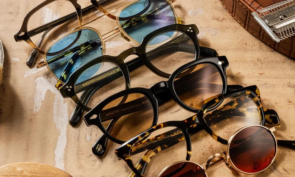 Multiple pairs of sunglasses with different frame styles and colors are placed on a textured surface, along with a small pen and a wooden box.