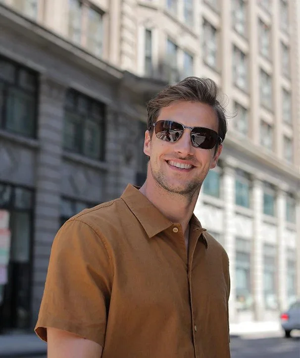 A smiling man wearing sunglasses and a brown short-sleeve button-up shirt standing on a city street.
