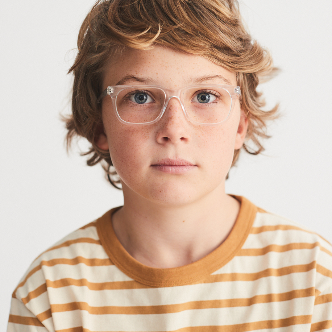 A young boy with curly blond hair and light blue eyes wearing clear glasses and a tan and white striped shirt, looking directly at the camera with a neutral expression.
