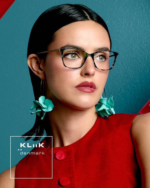 A woman with black hair, glasses, and large turquoise earrings poses against a teal background, wearing a red top with buttons.