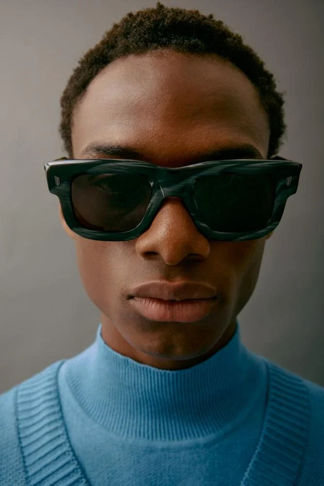Close-up of a person wearing large, dark sunglasses and a blue turtleneck sweater, with a plain gray background.