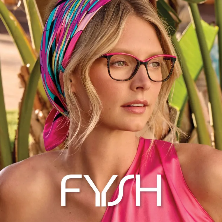 A woman wearing glasses and a colorful headscarf, with a pink top, standing outdoors with large green leaves in the background.