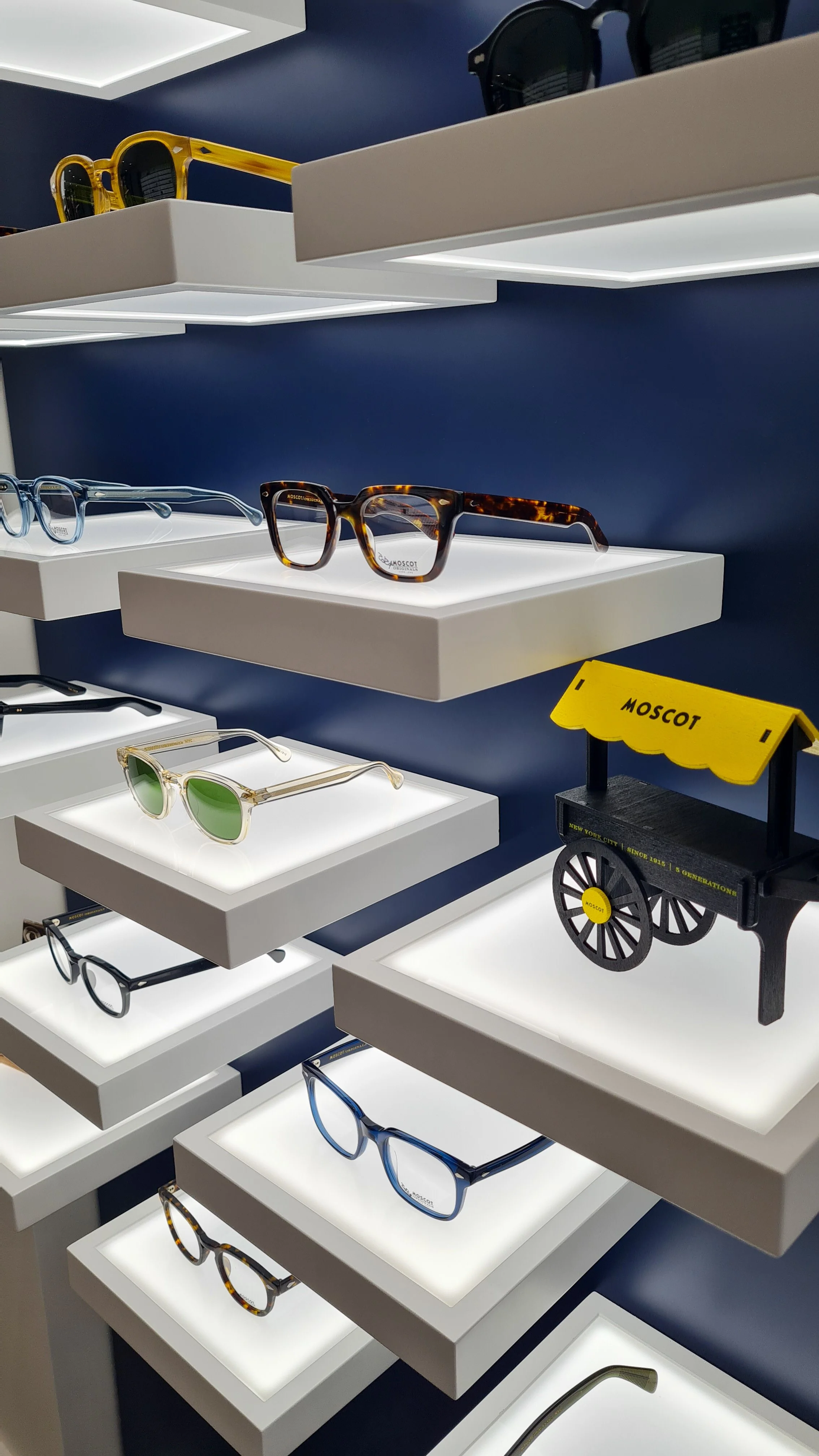 Display of various eyeglasses and sunglasses on illuminated shelves in a store, with a small decorative cart labeled "MOSCOT".