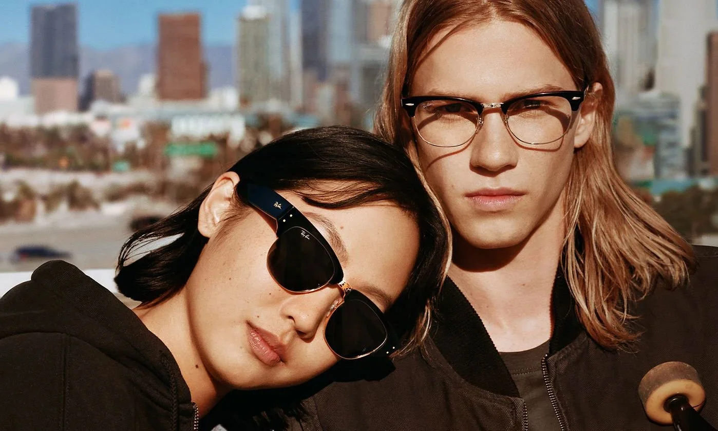 A young woman with short black hair wearing Ray-Ban sunglasses resting her head on a young man with shoulder-length red hair, glasses, and a black jacket, with an urban cityscape in the background.
