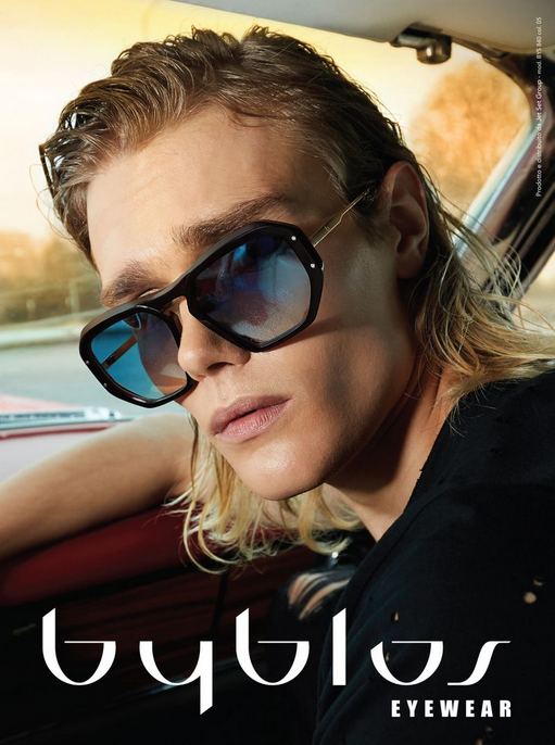 A woman with shoulder-length blond hair wearing black sunglasses with blue-tinted lenses, sitting in a car, looking out of the window.