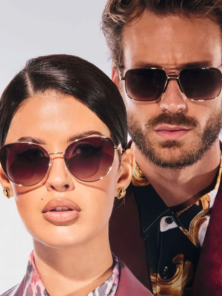 Close-up of a woman and a man wearing sunglasses, posing together against a plain background.