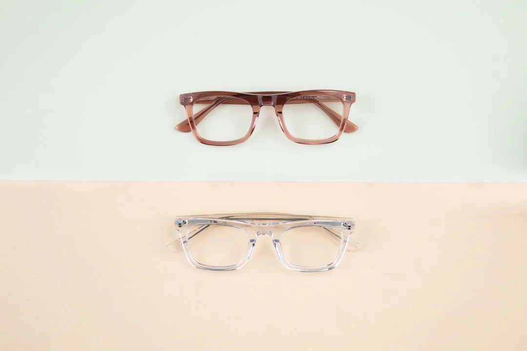 Two pairs of eyeglasses, one pink and the other transparent, placed on a table with a pastel-colored background.