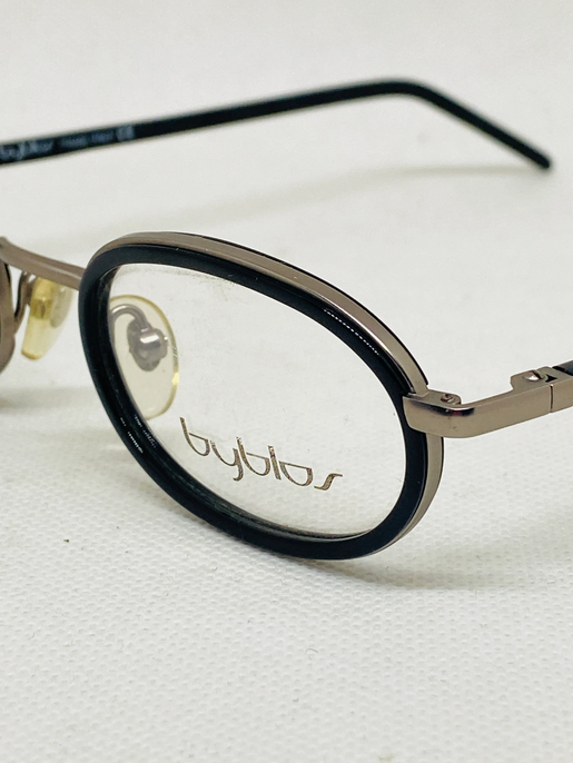 Close-up of a pair of eyeglasses with black and metallic frames, showing the brand name 'byblos' on the lens.