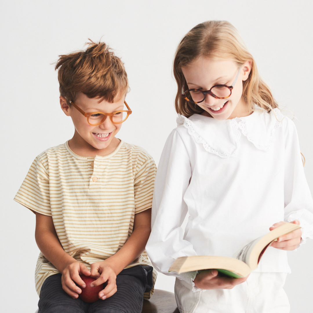 A young girl and a young boy wearing glasses sit together, smiling and looking at a book that the girl is holding, against a plain white background.