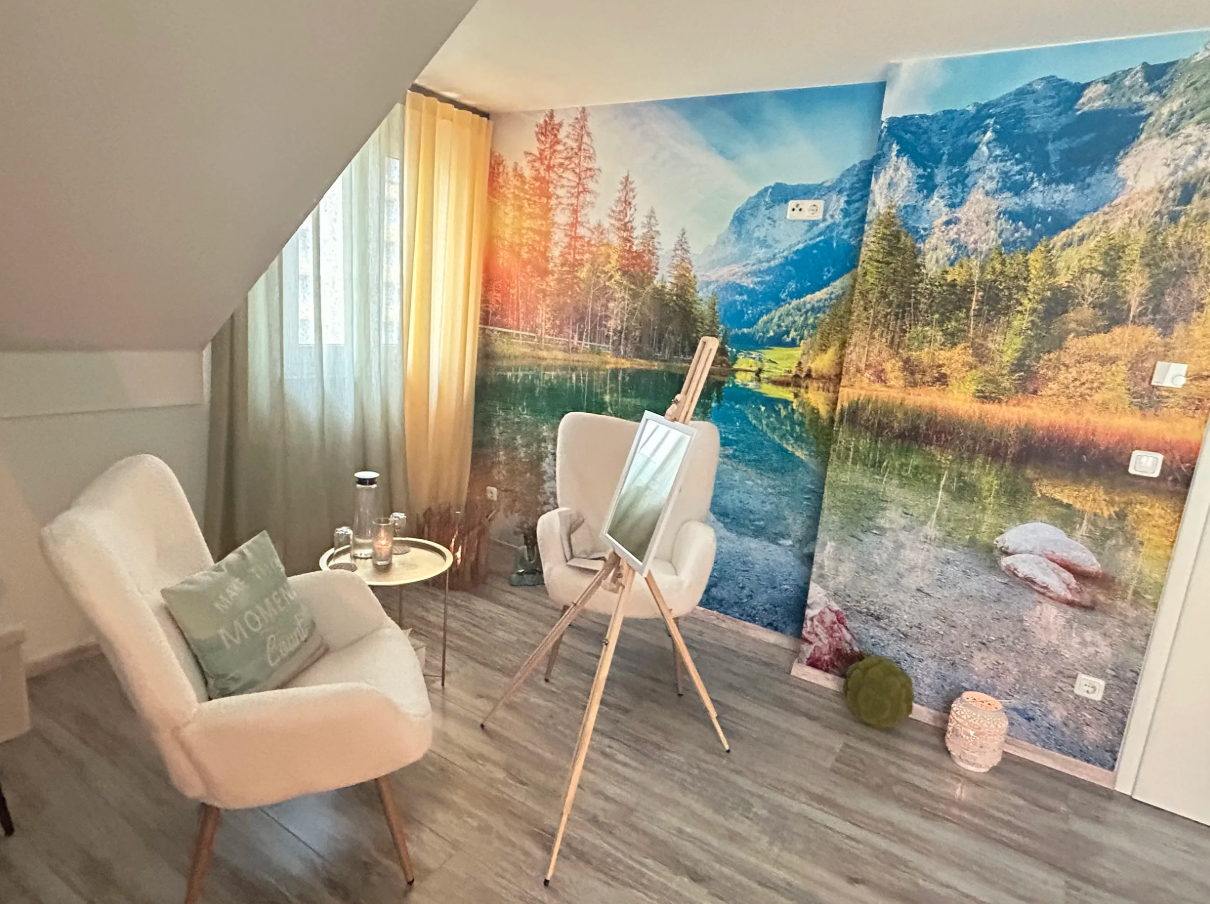 Ein gemütliches Wohnzimmer mit einer Natur-Fototapete, die einen See, Bäume und Berge zeigt. Es gibt zwei weiße Sessel, ein kleines Beistelltischchen, einen Spiegel auf einem Stativ, eine grüne Kissen und einige Dekorationsgegenstände auf dem Boden.