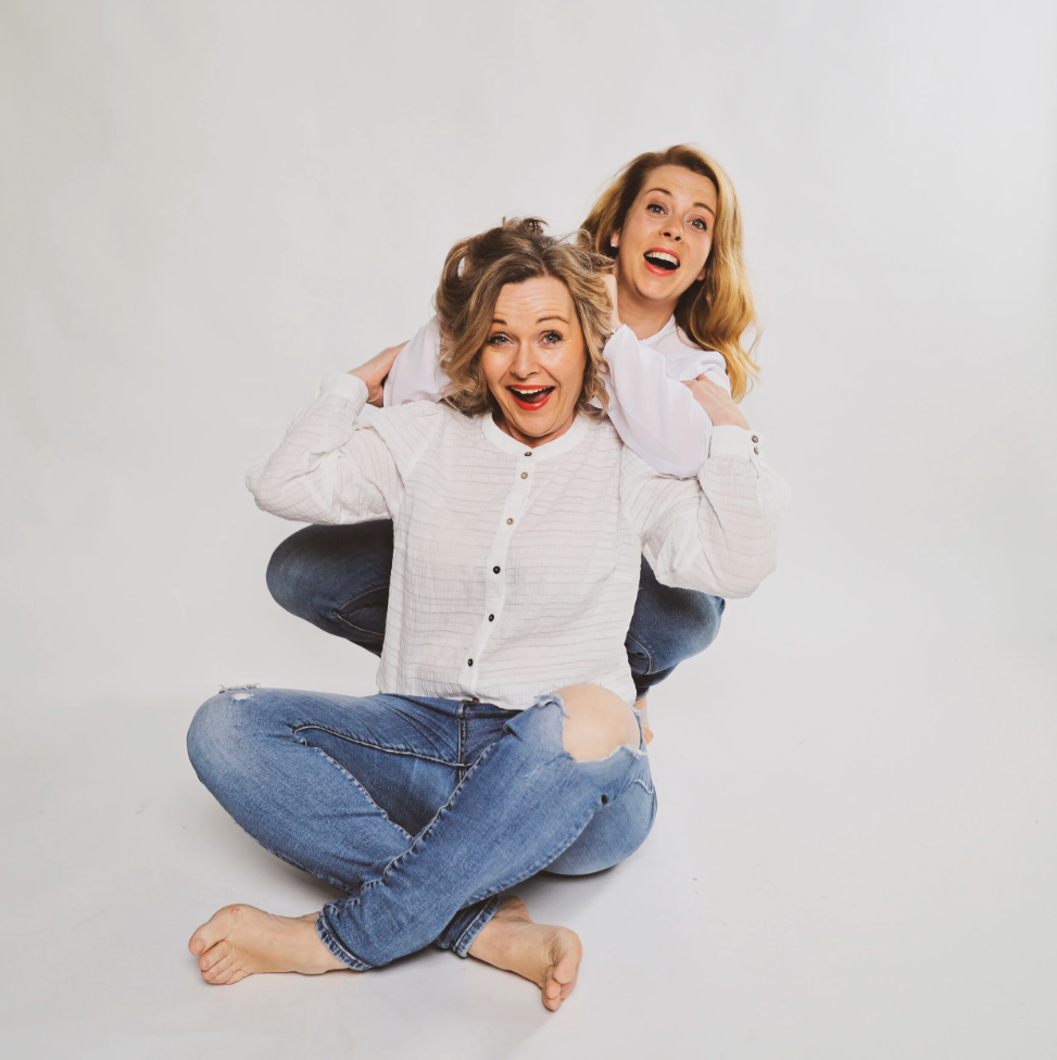 Zwei lachende Frauen, eine sitzt vor der anderen, die auf ihrem Rücken sitzt, beide tragen weiße Oberteile und Jeans und wirken fröhlich.