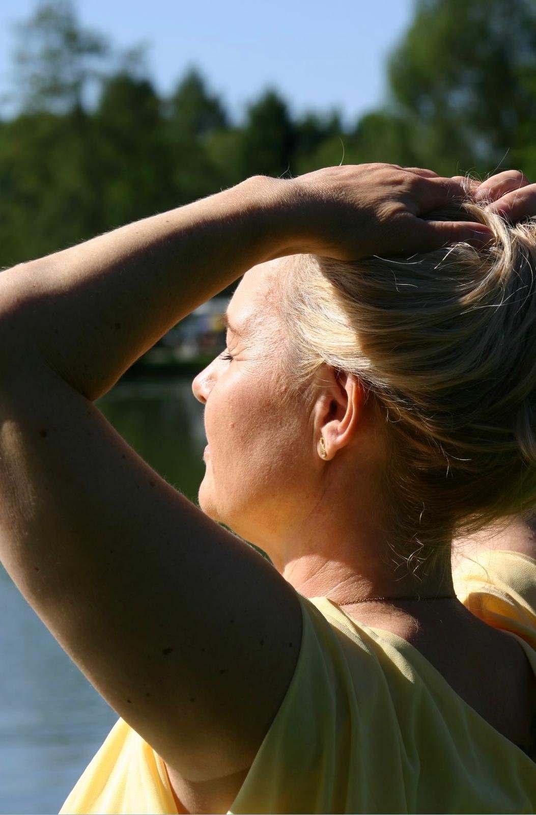 Eine Frau mit blonden Haaren trägt ein gelbes Oberteil und steht im Freien an einem Fluss, während sie die Augen schließt und ihre Hände im Haar hält. Der Himmel ist blau und die Bäume im Hintergrund sind grün.