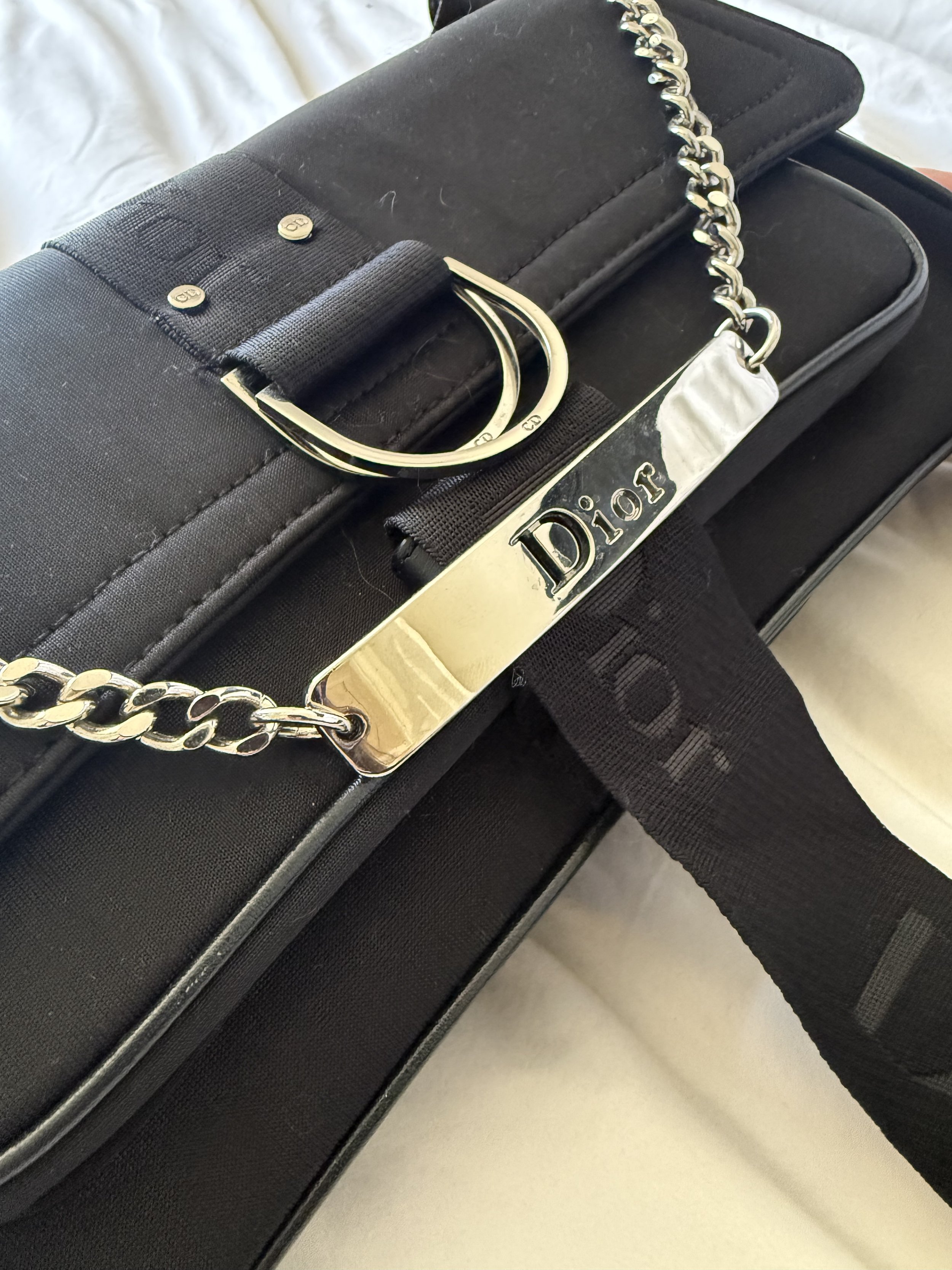 Close-up of a black Dior handbag with a silver chain strap and a tag, placed on a light-colored surface.