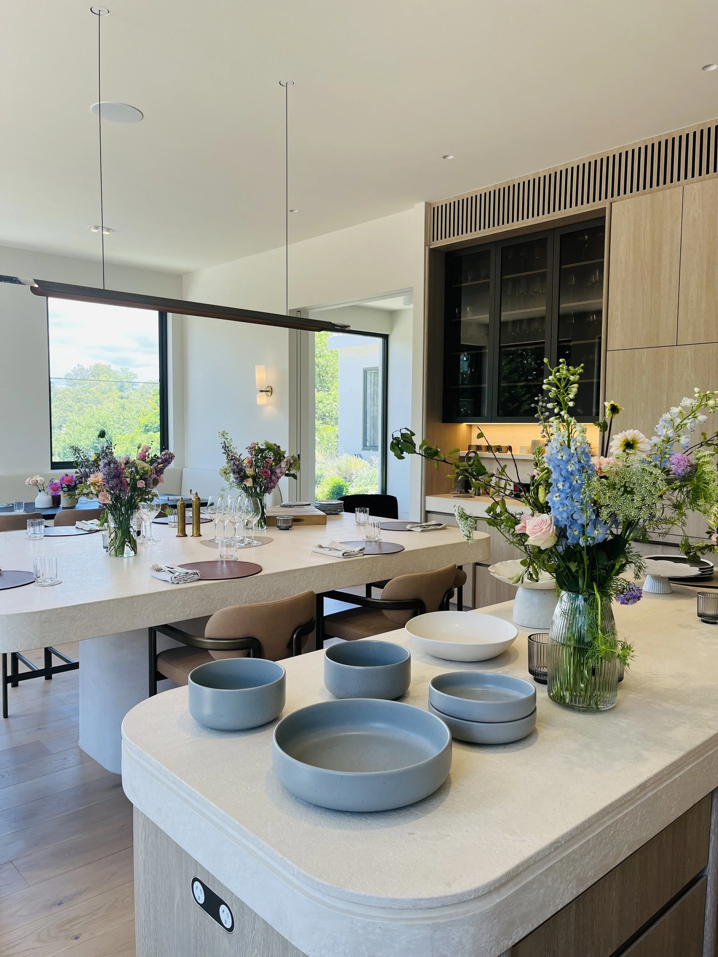 Modern dining room with a large table set with glasses, plates, and cutlery, decorated with floral centerpieces. Behind the table, there are large windows, and a kitchen area with light wood cabinets and black glass-fronted shelves. The room features contemporary lighting and a minimalistic design.