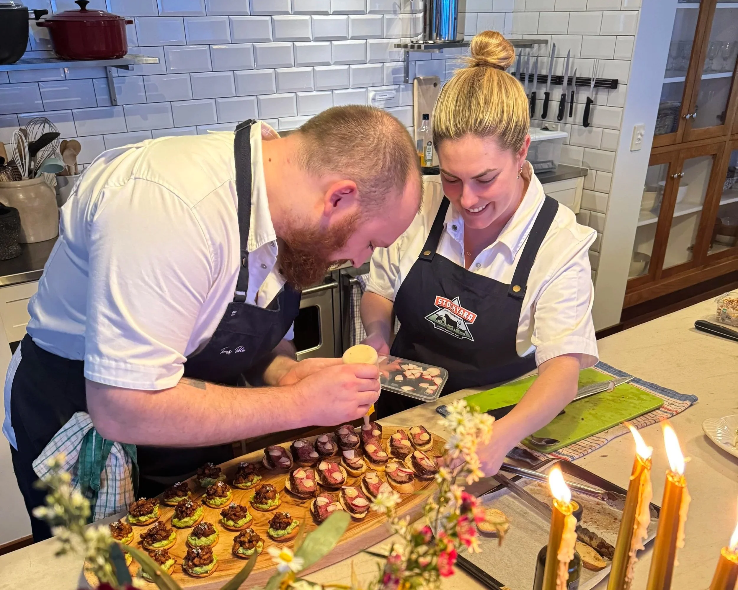 Two people in chef uniforms preparing and decorating various appetizers and finger foods on a kitchen counter, with a background of kitchen shelves and white tiled walls.