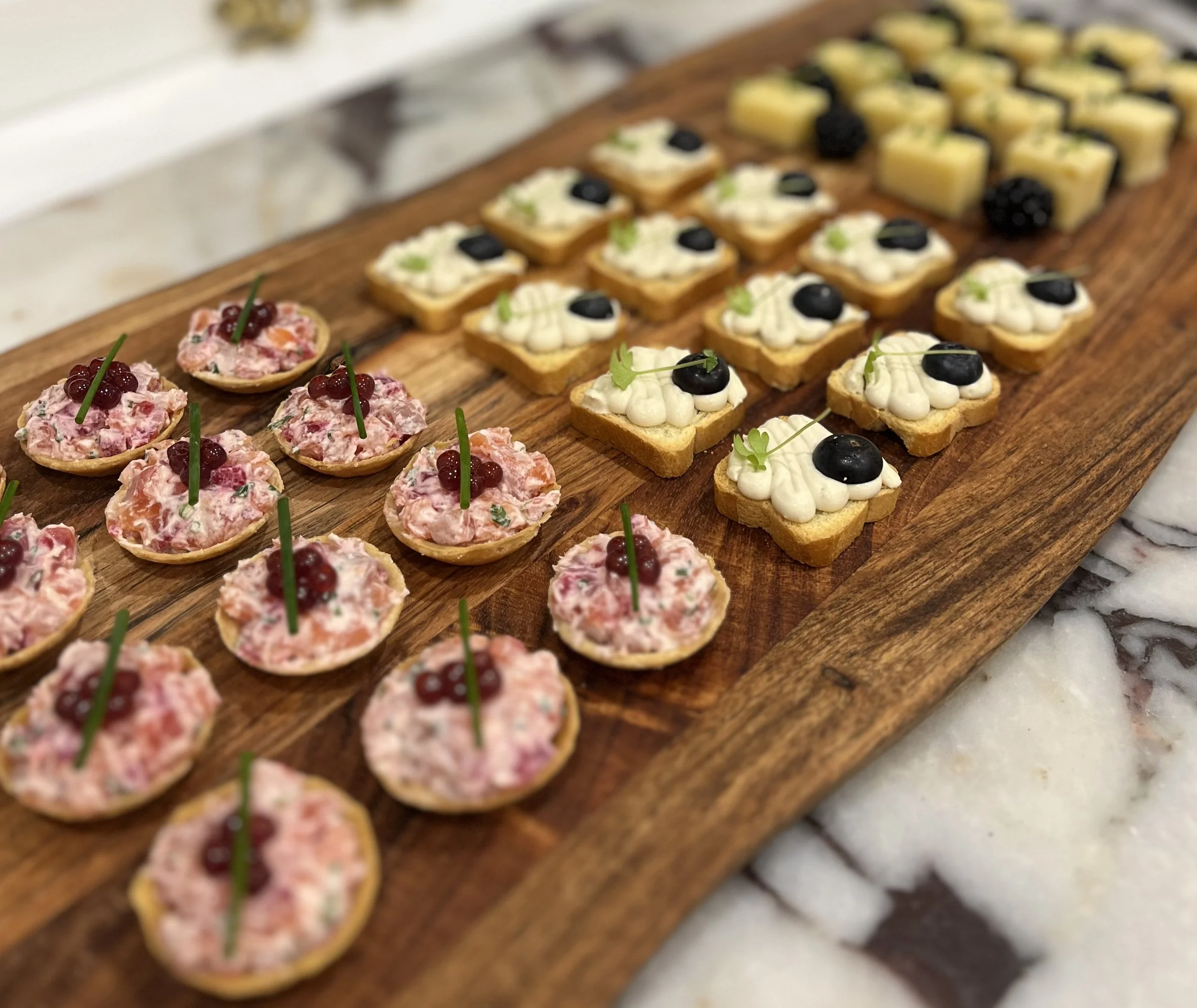 Assorted bite-sized appetizers on a wooden serving board, including tartlets with cream and caviar, and small square savory snacks with cheese and blackberries garnished with greens.