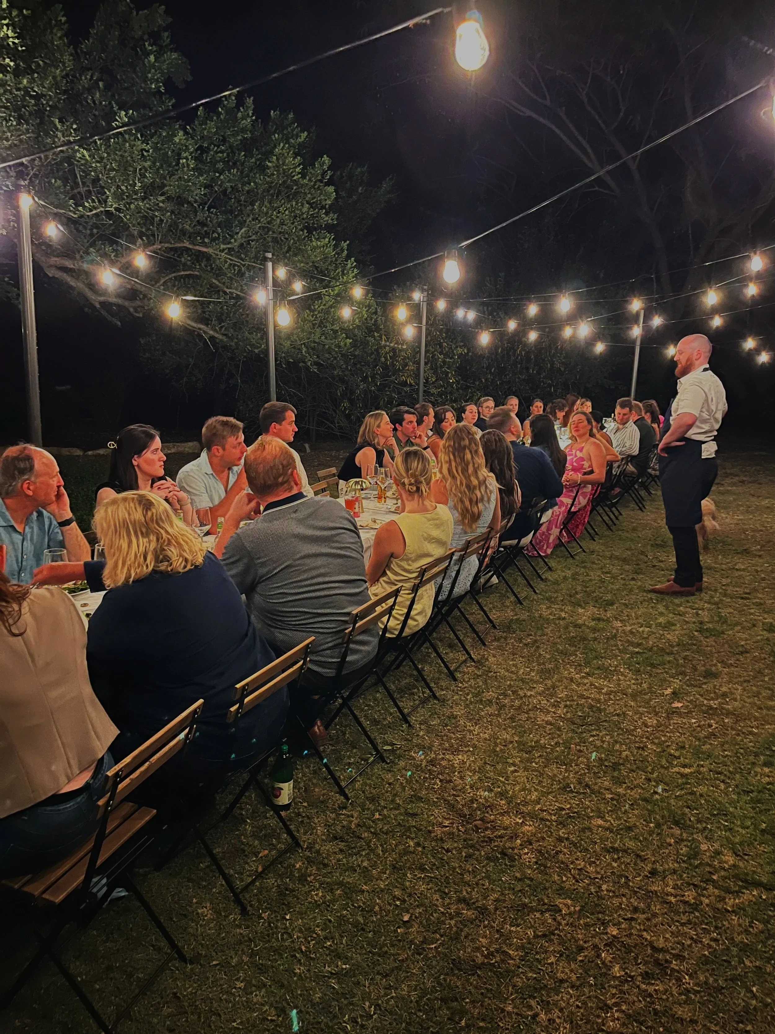 A group of people having dinner outdoors at night under string lights, with a man standing and speaking to the group.