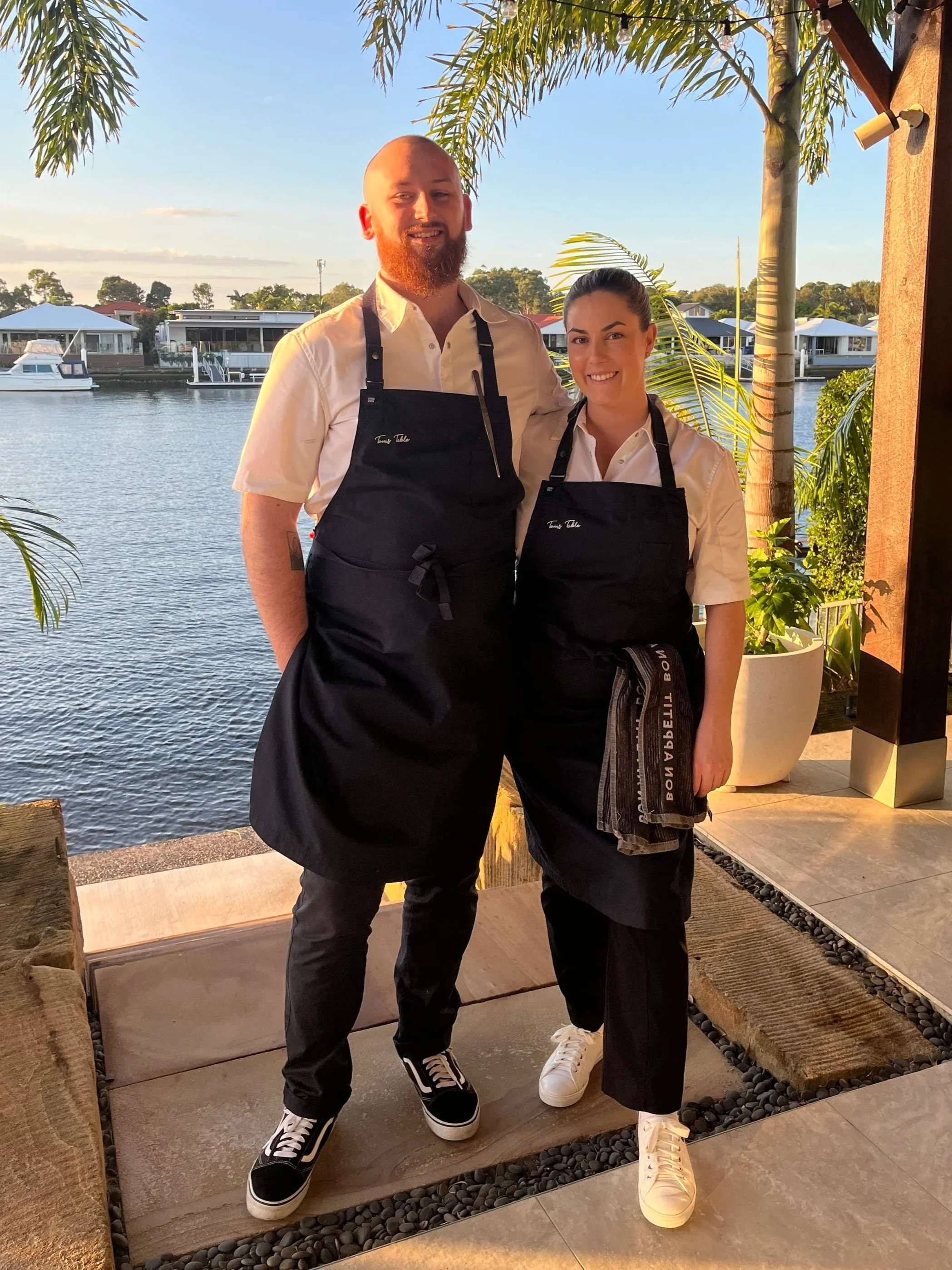 Two people, a man and a woman, standing side by side outdoors near water and boats, both wearing white shirts and black aprons, smiling for the photo.