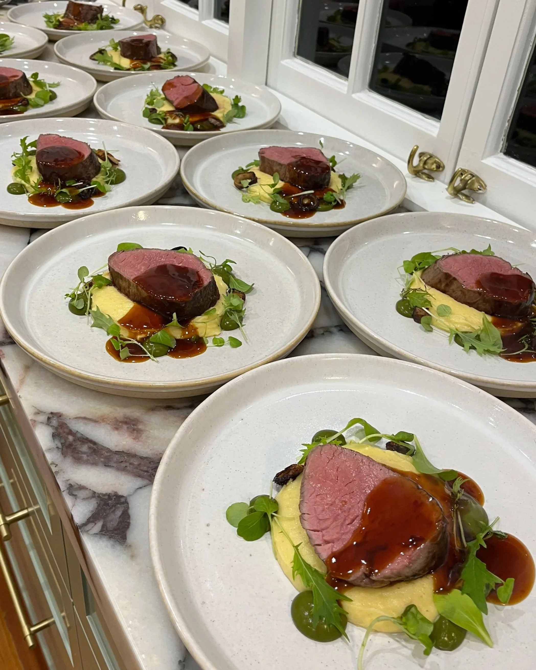 Close-up of plates with square-cut beef steak topped with sauce, served on a bed of mashed potatoes, garnished with green herbs and accompanied by green sauce on a white countertop near a window.