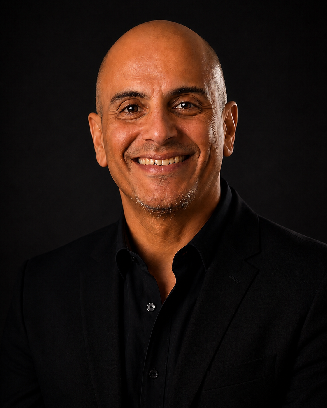 A smiling middle-aged man with a bald head, wearing a black suit and black shirt, against a dark background.