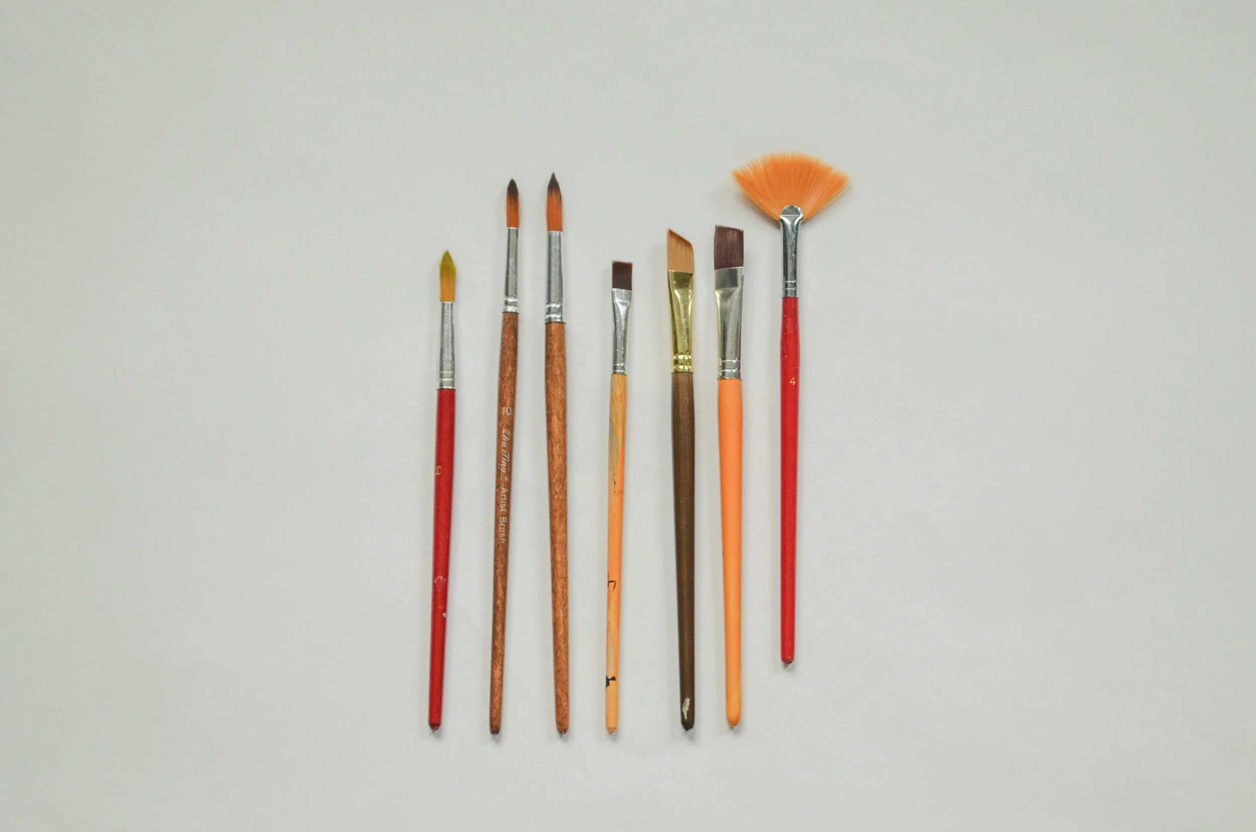 Eight small paintbrushes with different bristle shapes and sizes, arranged in a row on a white background.