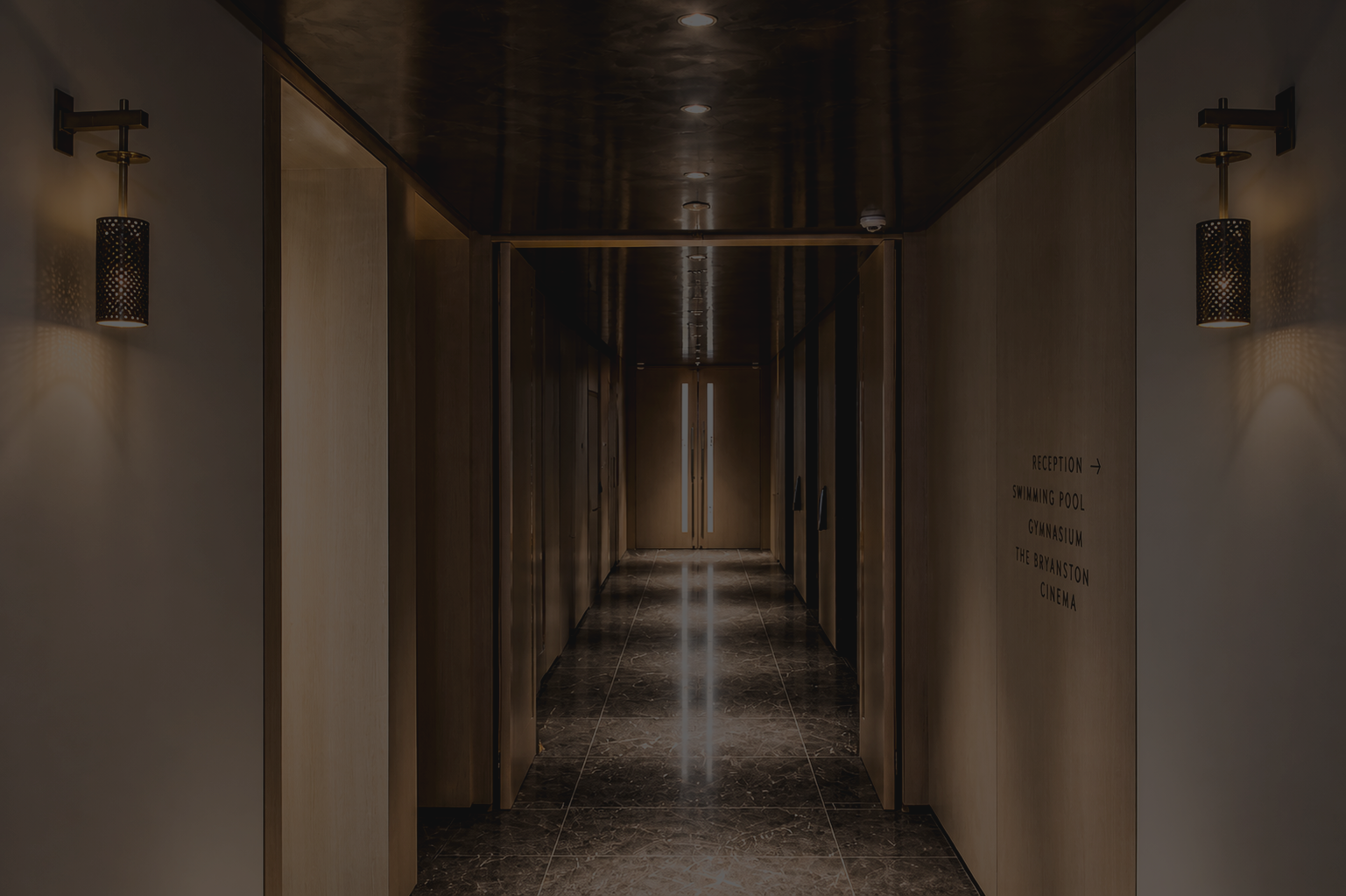 A dimly lit hallway with dark marble floors, wooden walls, and wall-mounted lamps, leading to a set of double doors at the end.