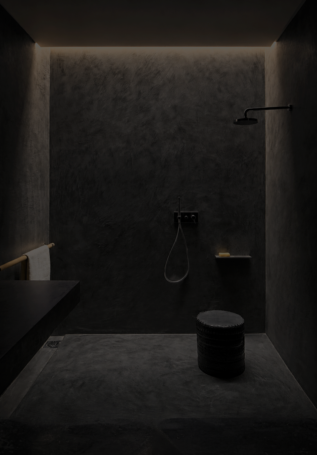 Minimalist black shower with a dark textured wall, ceiling light, black showerhead, handheld shower, small shelf with soap, black round stool, and towel on a railing.